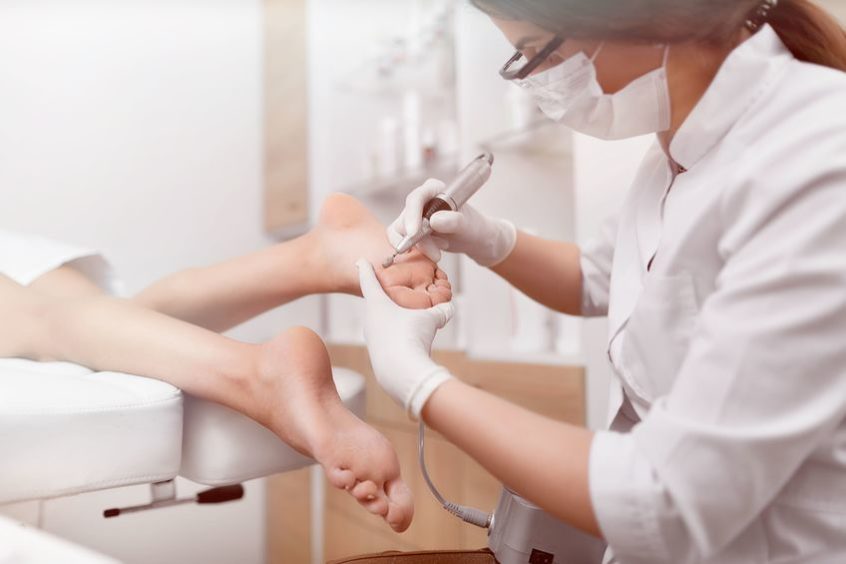 View of odiatrist using special grinding equipment and making procedure polish for feet. Podiatry specialist in white gloves cleaning skin of client from callus and corn with professional tools.
